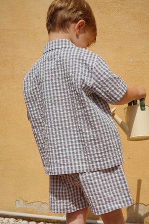 A young child stands sideways against a tan wall, holding a watering can and wearing the Konges Slojd Baby Boys’ and Boys’ Kim Short Sleeve Shirt in a classic blue and white checkered seersucker.