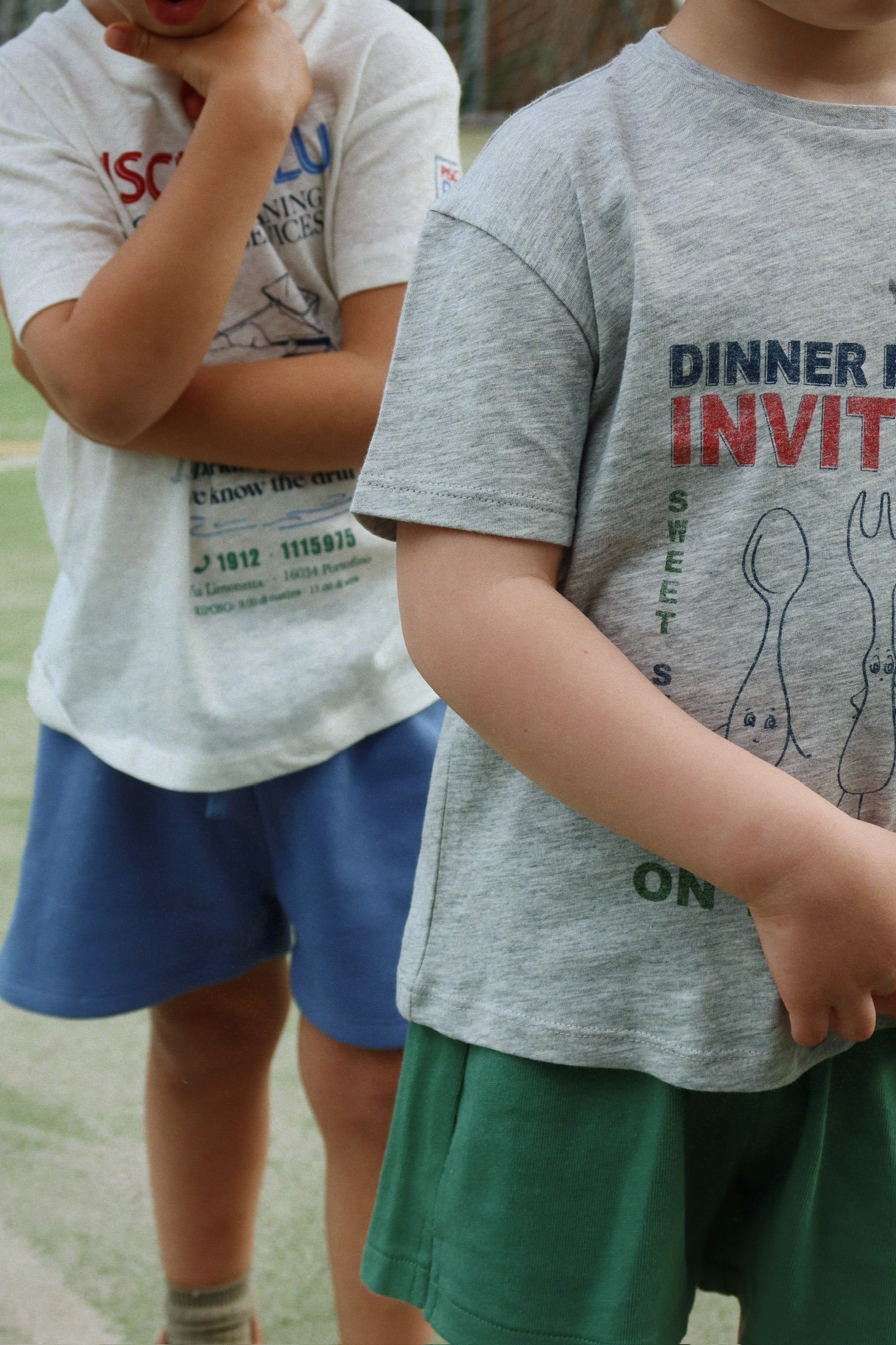 Two children stand outdoors on a green surface, both wearing Konges Slojd Baby Boys’ and Boys’ Spotty Sweat Shorts with an elastic waistband and t-shirts. One child has their arm across their chest while the other’s arms rest by their sides, with faces not visible.
