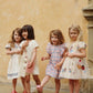 Four young girls in summer dresses and sandals stand together against a beige wall, appearing to pose for a photo—one of them wears the Konges Slojd Baby Girls’ and Girls’ Kim Frill Dress, adding a touch of effortless charm to the group’s breezy look.
