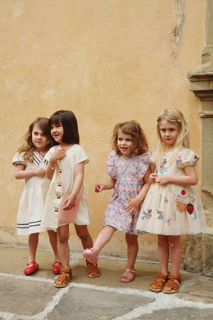 Four young girls in summer dresses and sandals stand together against a beige wall, appearing to pose for a photo—one of them wears the Konges Slojd Baby Girls’ and Girls’ Kim Frill Dress, adding a touch of effortless charm to the group’s breezy look.