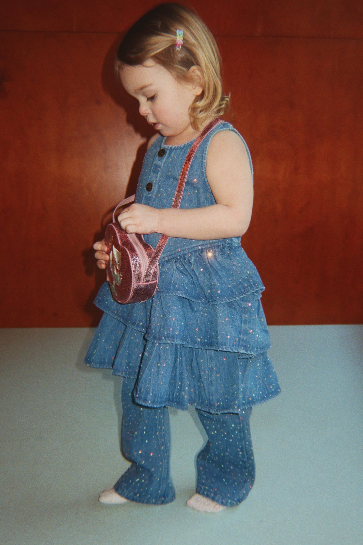 A toddler wearing a ruffled denim dress and matching Girls’ Elba Sparkle Jeans, holding a small pink circular bag.