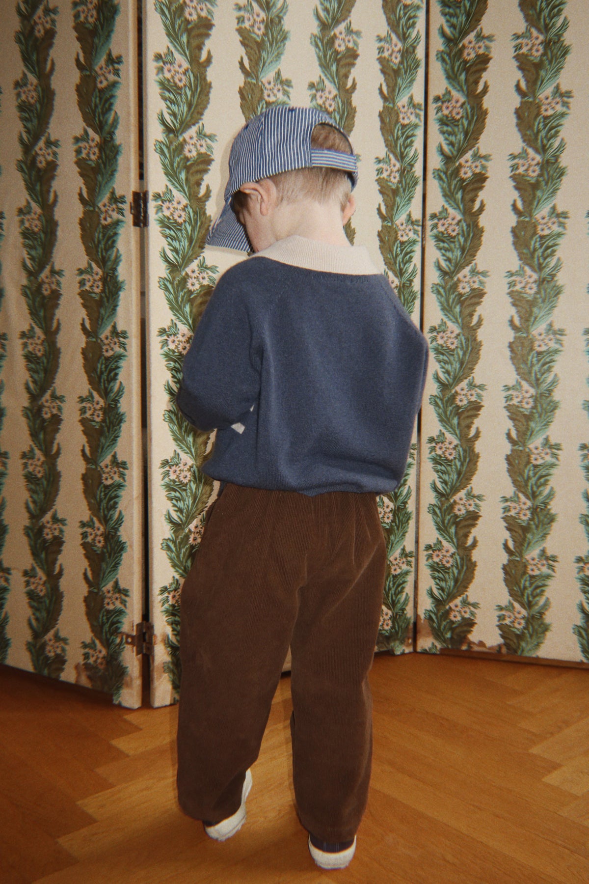 A child seen from behind, wearing a blue-striped cap, a blue long-sleeved shirt, and Konges Slojd Baby Boys’ and Boys’ Remy Pants in brown.