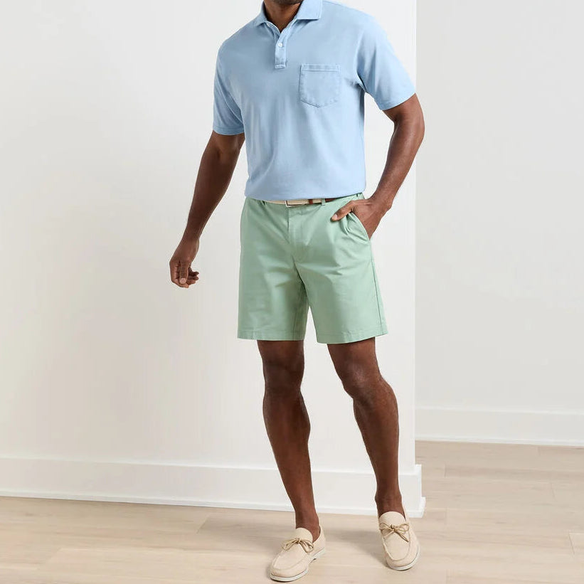 A man stands indoors wearing a light blue polo shirt, Peter Millar Crown Comfort Short in mint green with an 8” inseam and plain weave fabric, paired with a beige belt and beige loafers.