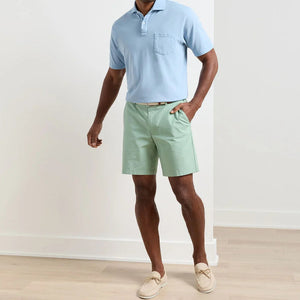 A man stands indoors wearing a light blue polo shirt, Peter Millar Crown Comfort Short in mint green with an 8” inseam and plain weave fabric, paired with a beige belt and beige loafers.
