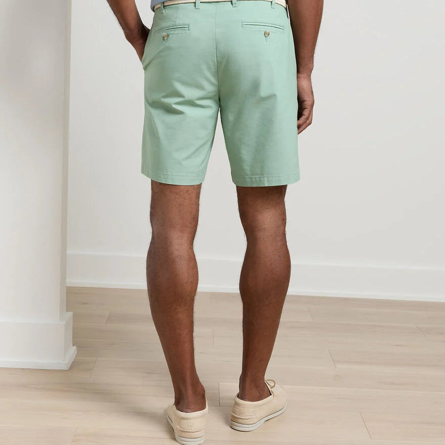 A man stands indoors facing away, wearing Peter Millar's Crown Comfort Short in light green with an 8" inseam and a white belt. The plain weave fabric is paired with beige loafers on a light wood floor against a white wall.