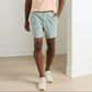 Indoors, a person wears a peach T-shirt and light green Peter Millar Crown Weekend Shorts with an elastic waist, paired with white sneakers on a light wood floor.
