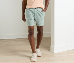 Indoors, a person wears a peach T-shirt and light green Peter Millar Crown Weekend Shorts with an elastic waist, paired with white sneakers on a light wood floor.