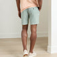 A person stands indoors facing away, wearing a peach t-shirt, Peter Millar Crown Weekend Short in light green stretch-cotton with a 7-inch inseam and back pocket, and white sneakers on a light wood floor.