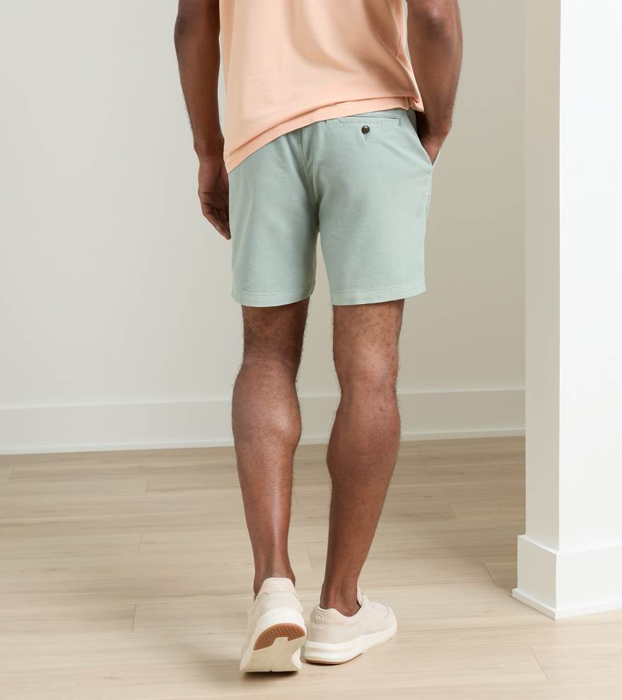 A person stands indoors facing away, wearing a peach t-shirt, Peter Millar Crown Weekend Short in light green stretch-cotton with a 7-inch inseam and back pocket, and white sneakers on a light wood floor.