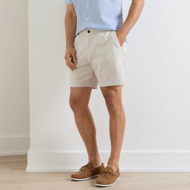 A man stands with hands in his pockets wearing Peter Millar Crown Weekend Shorts, a light blue short-sleeve shirt, and brown boat shoes.