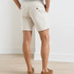 A man stands with hands in pockets, wearing Peter Millar Crown Weekend Short made from stretch-cotton, a light blue shirt, and brown boat shoes, facing away in a bright, minimal room.