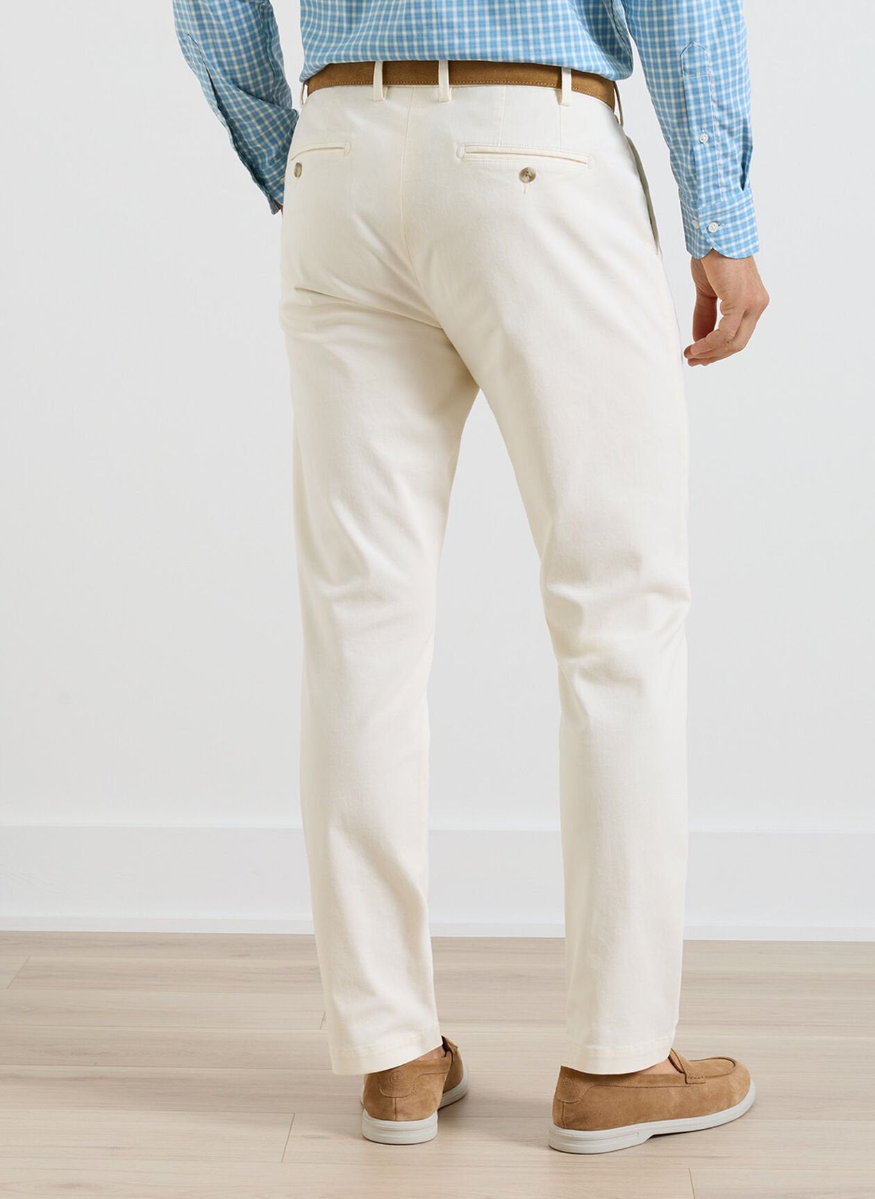 A man stands indoors facing away, wearing Peter Millar Signature Sateen Flat Front Trousers in cream, paired with a blue checkered shirt, brown belt, and tan shoes on a light wood floor.