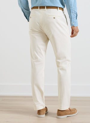 A man stands indoors facing away, wearing Peter Millar Signature Sateen Flat Front Trousers in cream, paired with a blue checkered shirt, brown belt, and tan shoes on a light wood floor.