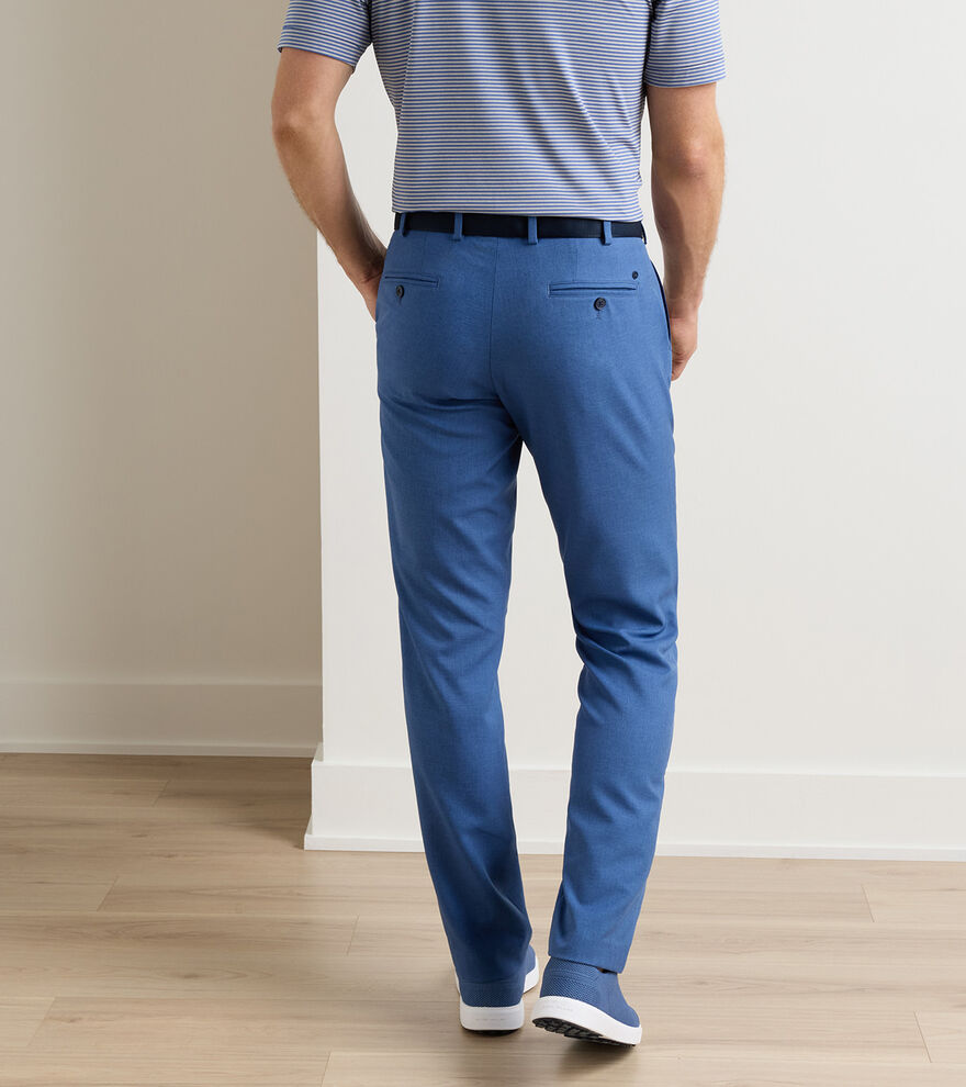 A man stands indoors on a light wood floor, seen from behind, wearing the Peter Millar Charlotte Performance Trouser with a blue striped polo and modern blue slip-on shoes.