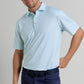 A man wearing a light blue Ashgrove Performance Jersey Polo by Peter Millar with UPF 50+ and dark blue pants stands with one hand on his hip against a plain background.