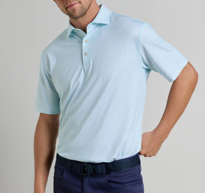 A man wearing a light blue Ashgrove Performance Jersey Polo by Peter Millar with UPF 50+ and dark blue pants stands with one hand on his hip against a plain background.
