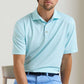 A man wearing the Peter Millar Dunnes Performance Jersey Polo—light blue, short-sleeve, collared, with thin white stripes and four-way stretch—paired with light blue pants, sits on a chair.