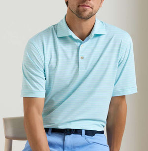 A man wearing the Peter Millar Dunnes Performance Jersey Polo—light blue, short-sleeve, collared, with thin white stripes and four-way stretch—paired with light blue pants, sits on a chair.