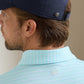 A man is shown from the back wearing a Peter Millar Dunnes Performance Jersey Polo, a light blue striped collared shirt made from four-way stretch fabric, paired with a navy cap.