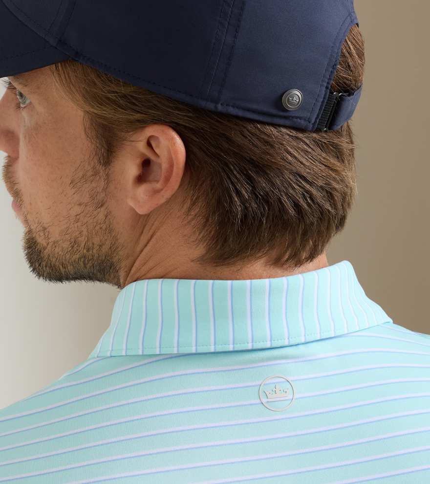 A man is shown from the back wearing a Peter Millar Dunnes Performance Jersey Polo, a light blue striped collared shirt made from four-way stretch fabric, paired with a navy cap.