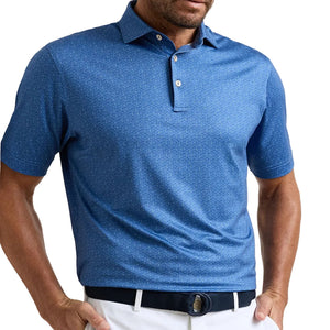 A man stands indoors, hands on hips and looking down, wearing a Peter Millar Negroni Performance Jersey Polo in blue with UPF 50+ protection, paired with white pants and a black belt.