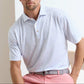 A man sits on a chair wearing a light blue patterned Peter Millar Nice Shot Performance Jersey Polo with four-way stretch, paired with pink pants and a grey belt against a plain background.