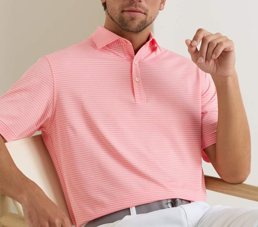 A man sits on a light chair wearing the Peter Millar Merion Performance Jersey Polo in pink and white stripes with UPF 50+ protection and light pants.