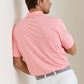 A man wearing a Peter Millar Merion Performance Jersey Polo in light pink and white stripes with four-way stretch, paired with white pants, stands with his back to the camera, leaning against a wall.