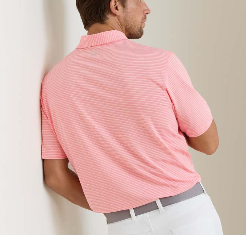 A man wearing a Peter Millar Merion Performance Jersey Polo in light pink and white stripes with four-way stretch, paired with white pants, stands with his back to the camera, leaning against a wall.