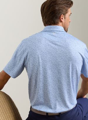 A man with brown hair, shown from the back on a wicker chair against a plain background, wears the Peter Millar Parker Performance Jersey Polo in light blue with short sleeves and four-way stretch.