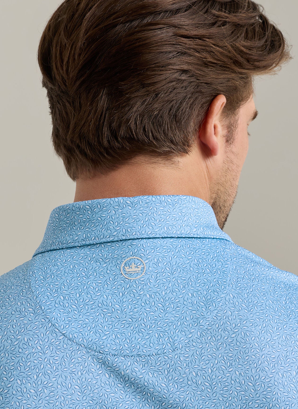 A man is shown from behind wearing the Peter Millar Magnolia Performance Jersey Polo in light blue with a pattern, four-way stretch, and a small white embroidered logo at the neck.
