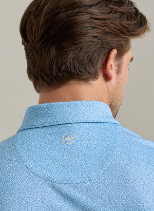 A man is shown from behind wearing the Peter Millar Magnolia Performance Jersey Polo in light blue with a pattern, four-way stretch, and a small white embroidered logo at the neck.