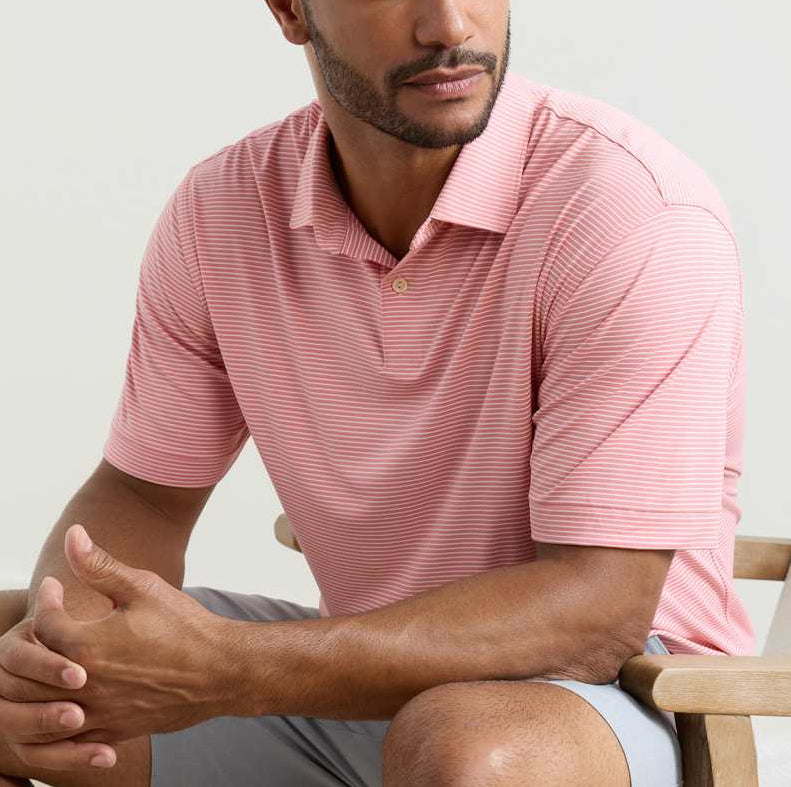 A man seated on a chair wears the Peter Millar Featherweight Stripe Performance Polo in light pink and white, paired with light gray shorts.