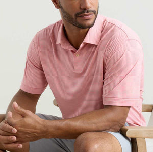 A man seated on a chair wears the Peter Millar Featherweight Stripe Performance Polo in light pink and white, paired with light gray shorts.