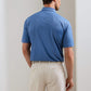 A man stands facing away in a blue Peter Millar Featherweight Headcovers Performance Polo with four-way stretch and light trousers, in a neutral indoor setting.
