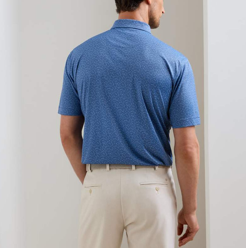 A man stands facing away in a blue Peter Millar Featherweight Headcovers Performance Polo with four-way stretch and light trousers, in a neutral indoor setting.