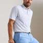 A man wears the Peter Millar Featherweight Cornhole Performance Polo, a short-sleeve, lightweight shirt with UPF 50+ sun protection, tucked into light blue pants with a grey belt, standing against a plain light background.