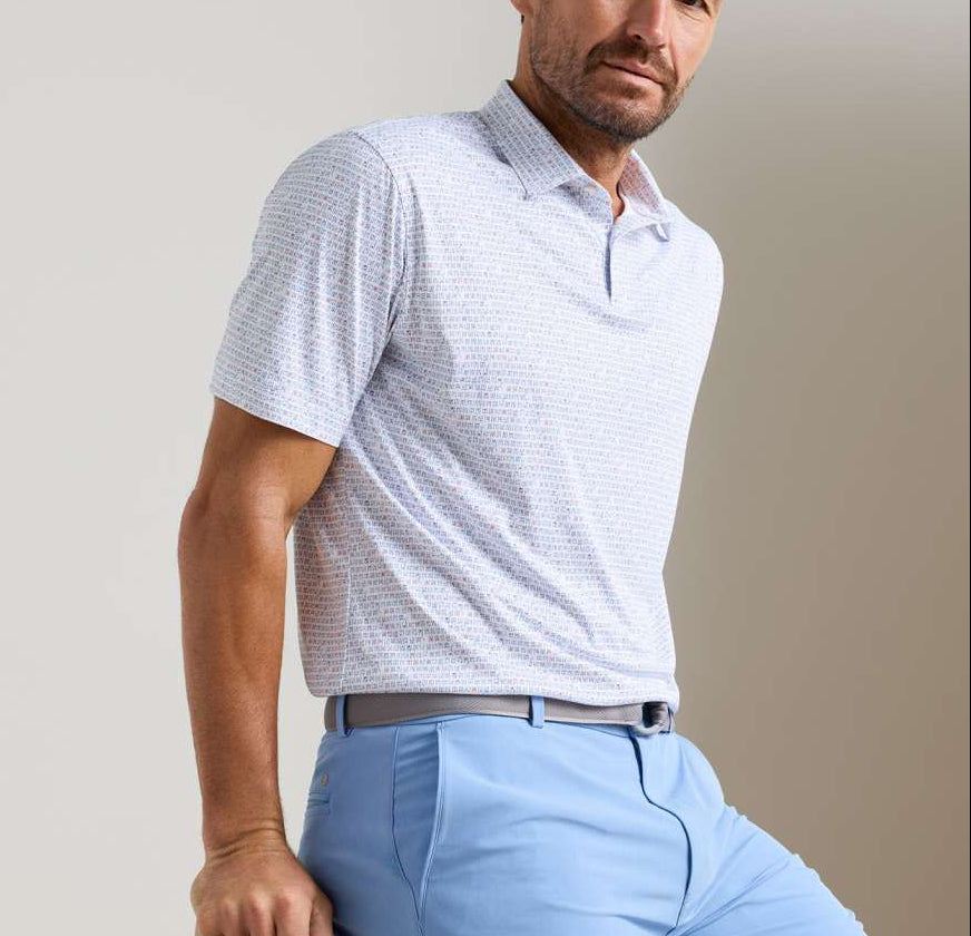 A man wears the Peter Millar Featherweight Cornhole Performance Polo, a short-sleeve, lightweight shirt with UPF 50+ sun protection, tucked into light blue pants with a grey belt, standing against a plain light background.