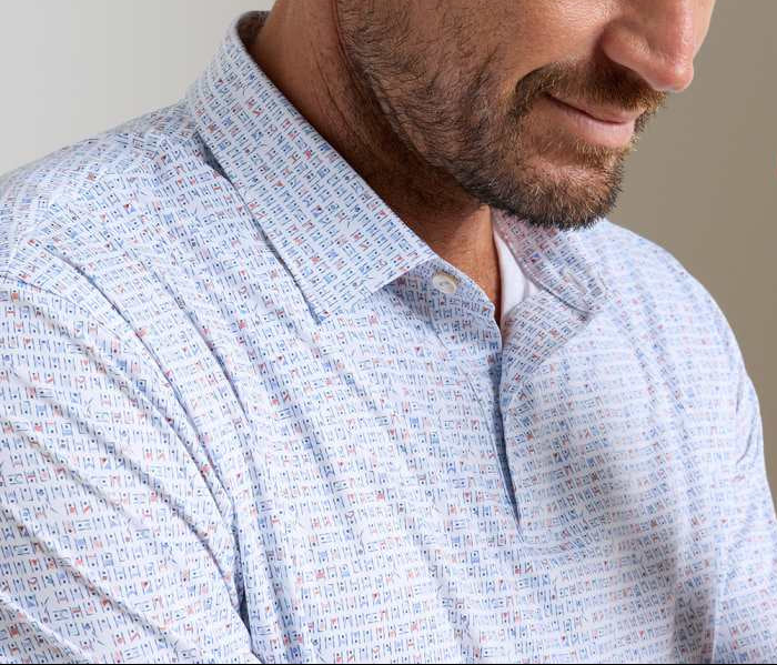 A man wears the Peter Millar Featherweight Cornhole Performance Polo—a light button-up with a multicolored geometric print and four-way stretch, shown from the shoulders up for a polished, all-day comfortable look.