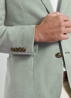 A person is shown from the chest down adjusting the front of a light green, garment-dyed Weekend Soft Jacket by Peter Millar, featuring brown buttons.