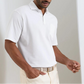 A man stands indoors in Peter Millar’s Everyday Pique Polo—white cotton piqué with a spread collar—paired with light pants for a look that combines classic style and lived-in softness.
