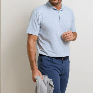 A man wears the Peter Millar Calvert Crown Flex Cotton Polo in light blue stripes and blue pants, holding a gray garment against a plain light wall—showcasing refined casual style.