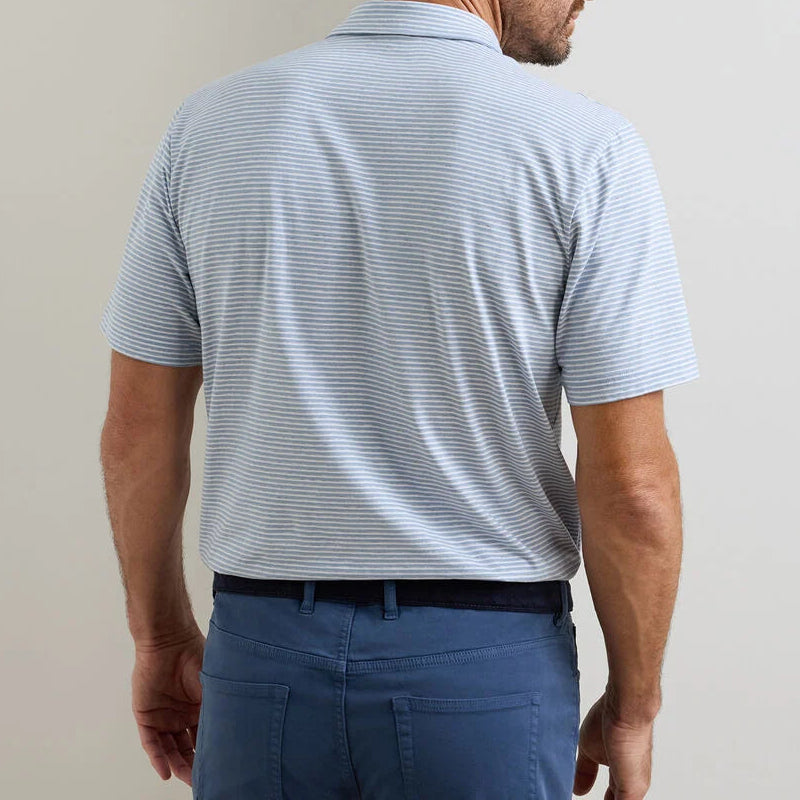 A man faces away in a Peter Millar Calvert Crown Flex Cotton Polo—light blue and white striped with a cutaway collar—paired with blue pants, showcasing timeless casual style against a plain backdrop.