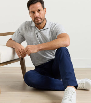 A man in a light gray Peter Millar Gilman Crown Flex Cotton Polo and navy pants sits on the floor beside a chair, looking at the camera.