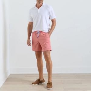 A man stands indoors against a plain white wall wearing Peter Millar’s Crown Swim Trunk in pink, featuring an elastic drawstring waistband and mesh liner, paired with a white polo shirt and brown boat shoes.