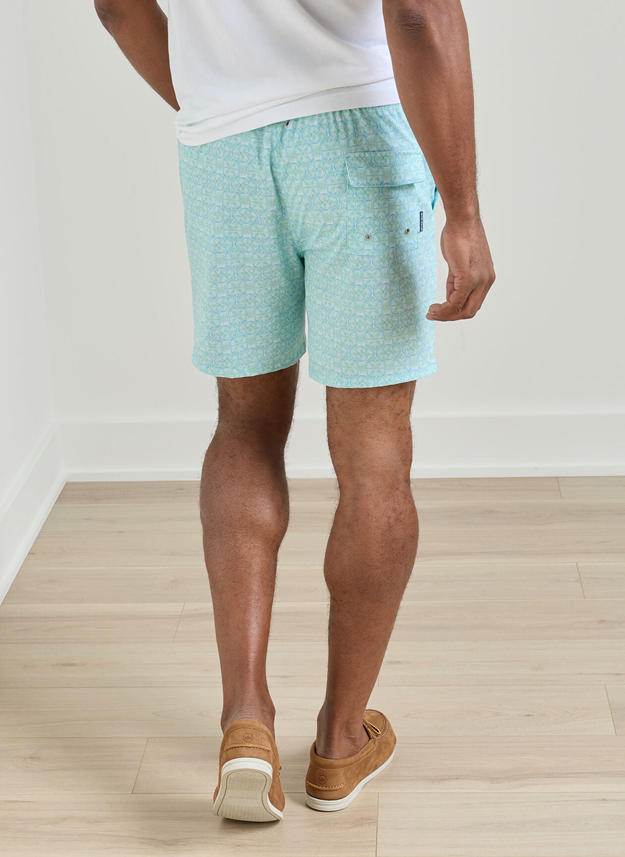 A person stands on a light wood floor facing away from the camera, wearing Peter Millar Tide Lines Swim Trunk in light blue with an elastic drawstring waistband, paired with a white shirt and brown loafers.