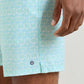 A close-up of a hand near light blue patterned Tide Lines Swim Trunks by Peter Millar, showing a small round logo by the hem and an elastic drawstring waistband for adjustable comfort.