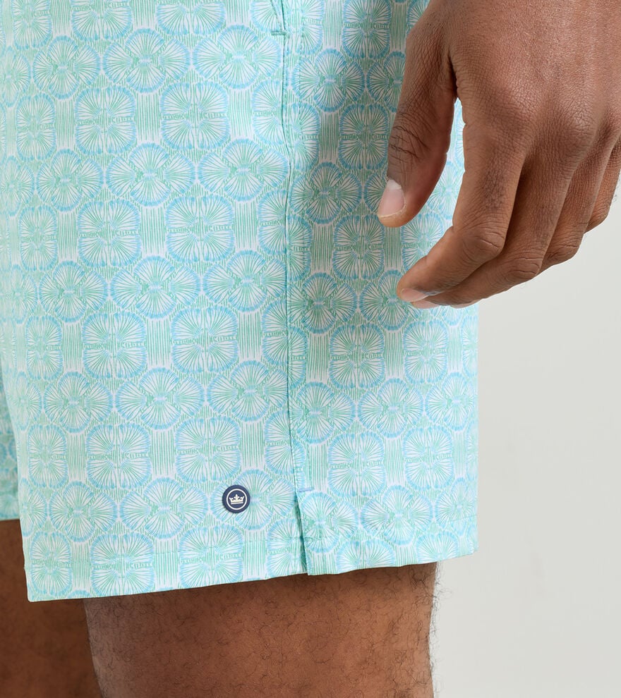 A close-up of a hand near light blue patterned Tide Lines Swim Trunks by Peter Millar, showing a small round logo by the hem and an elastic drawstring waistband for adjustable comfort.
