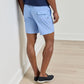 A man stands indoors facing away from the camera, wearing Peter Millar’s Nautical Ropes Swim Trunk—patterned light blue quick-drying swim trunks with an elastic waistband—paired with a navy shirt and navy slip-on shoes.