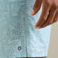 A close-up of a hand rests beside Peter Millar’s Seaside Shade Swim Trunk, featuring a light blue leaf pattern, small circular logo, and an elastic drawstring waistband for adjustable comfort.